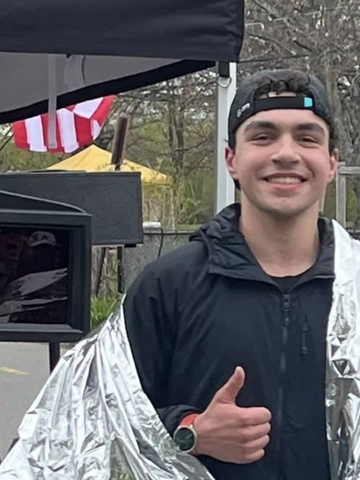 Alex Rogul at the Charles River Marathon finish line wrapped in a silver finisher blanket, giving a thumbs up.