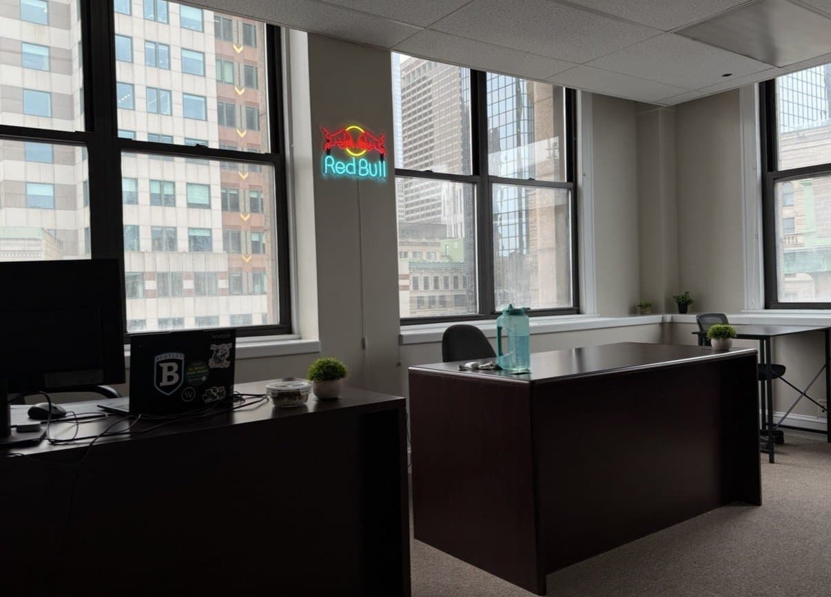 Corner office with tall windows onto downtown Boston towers, two dark wood desks, a laptop with a Bentley sticker, and a Red Bull neon sign on the wall.