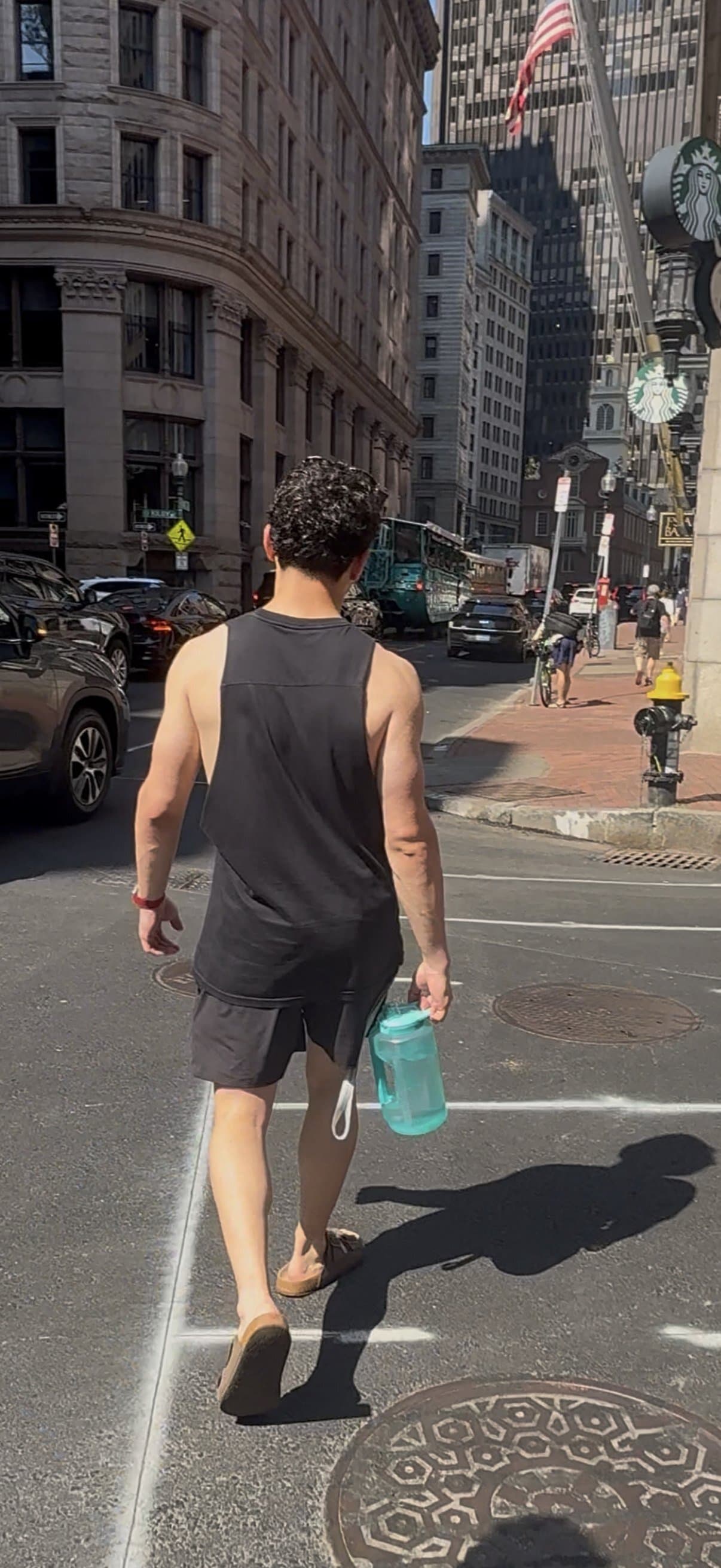 Alex seen from behind crossing a downtown Boston street in a black tank top, carrying a teal water jug, with granite office buildings ahead.