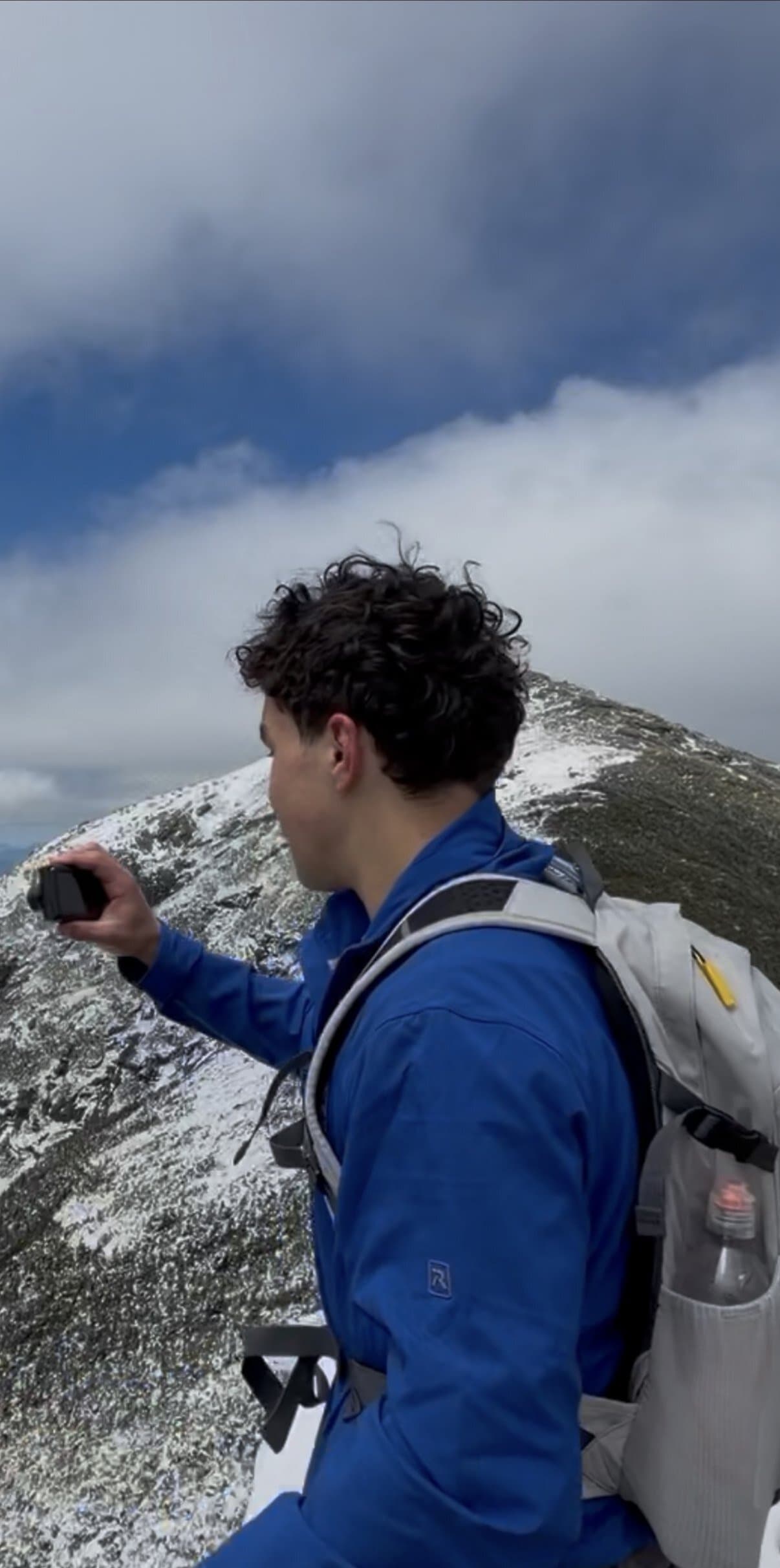 Alex in a blue jacket and grey pack on a snow-dusted mountain ridge, holding up a camera to film as clouds roll over the summit.